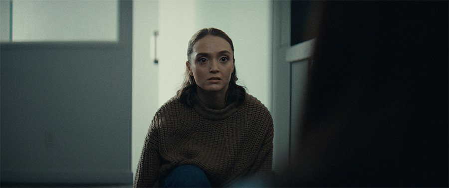 A woman wearing a brown sweater sits near a doorway