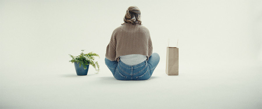 A woman sits on the floor between a fern and a paper bag