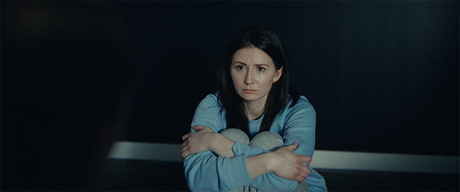 A woman in blue sits with arms wrapped around herself.